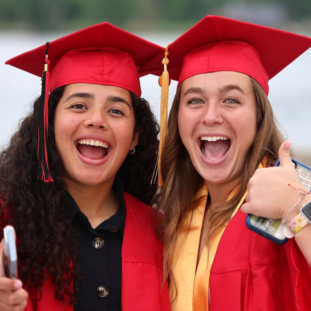 two female graduates standing in red cap and gown, smiling with thumbs up