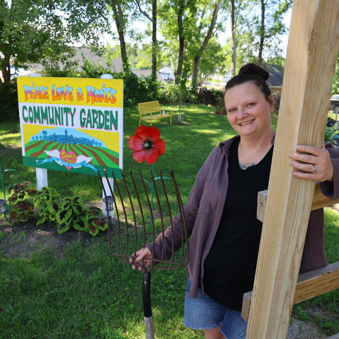 sarina standing in garden with tool in hand nex to green yellow sign for garden entrance